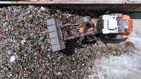 Drone-aerial-view-from-warehouse-glass-recycling-center,-following-forklift-excavator-grabbing-glass,-glass-recycling-concept