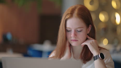 thoughtful brooding remote working red haired woman sitting infront of a laptop or notebook in casual outfit on her work desk in her modern airy bright living room home office with many windows