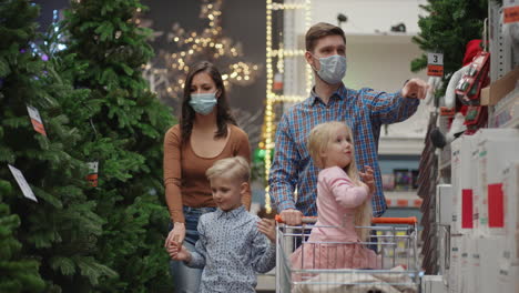 Una-Familia-De-Cuatro-Personas,-Madre,-Padre-E-Hijos,-Eligen-Juntos-Un-árbol-De-Navidad.-Una-Familia-Feliz-Con-Máscaras-Médicas-En-La-Tienda-Compra-Adornos-Navideños-Y-Regalos-En-Cámara-Lenta