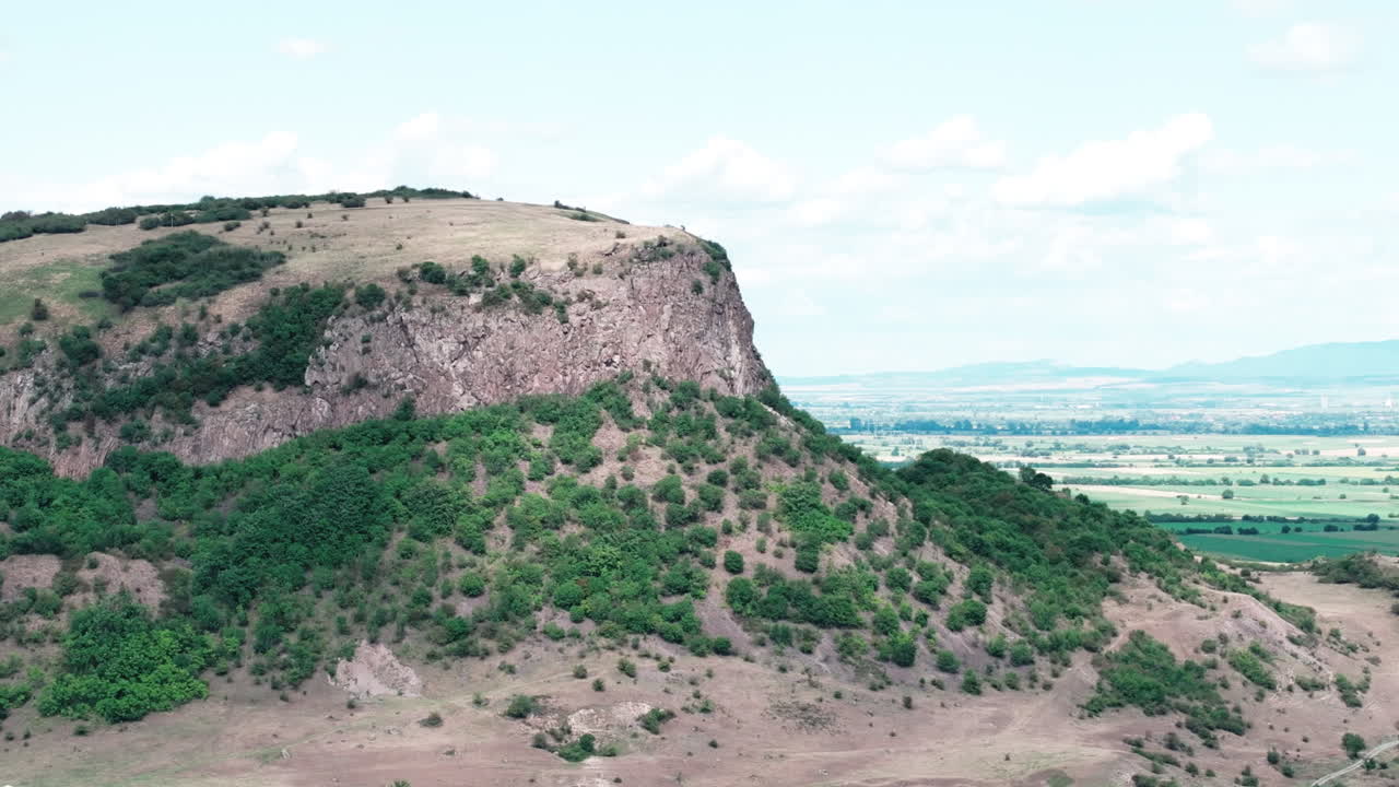 A majestic aerial drone shot orbits the Măgura Uroiului mountain in Romania. The view showcases the sheer rock cliff, green slopes, and the vast Transylvanian plain below