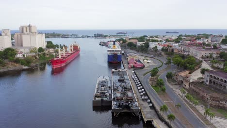 flight above ozama river by seaside road traffic towards large ships in downtown harbor port by blue ocean sea waters and sky, dominican republic, aerial approach