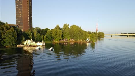 Parque-De-Wakeboard-Con-Un-Viejo-Barco-Frente-Al-Edificio-De-Televisión-De-Aspecto-Soviético-Con-Una-Torre-De-Televisión-En-Un-Fondo-Lejano-Junto-Al-Río-Daugava