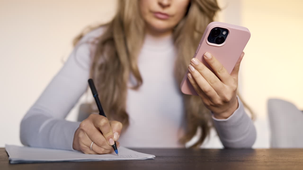 Woman writing on a piece of paper on a table while holding a phone