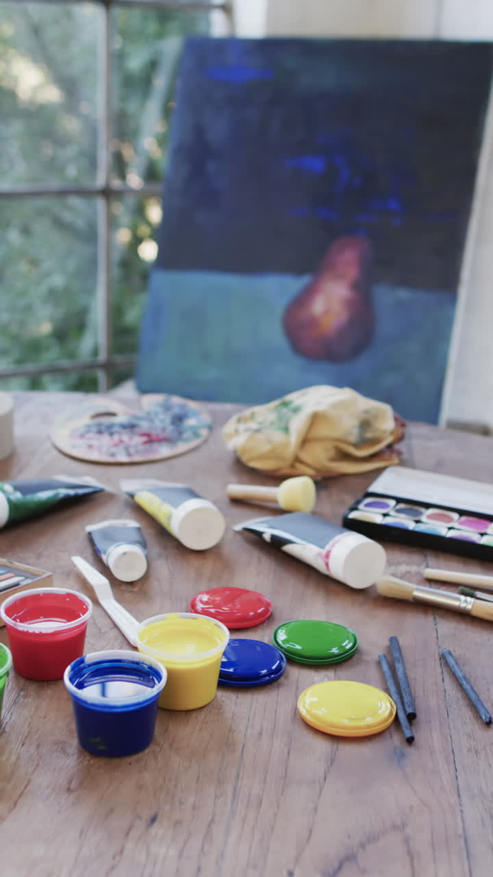 Vertical video of close up of tubes and tubs of paint and brushes on table in studio, slow motion