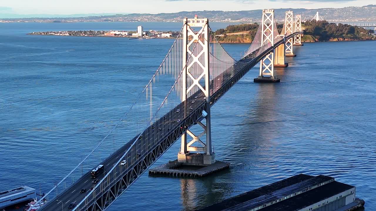 Drone captures a head-on view of the Bay Bridge with cars approaching, then arcs right as sunset light reflects off the bridge and bay
