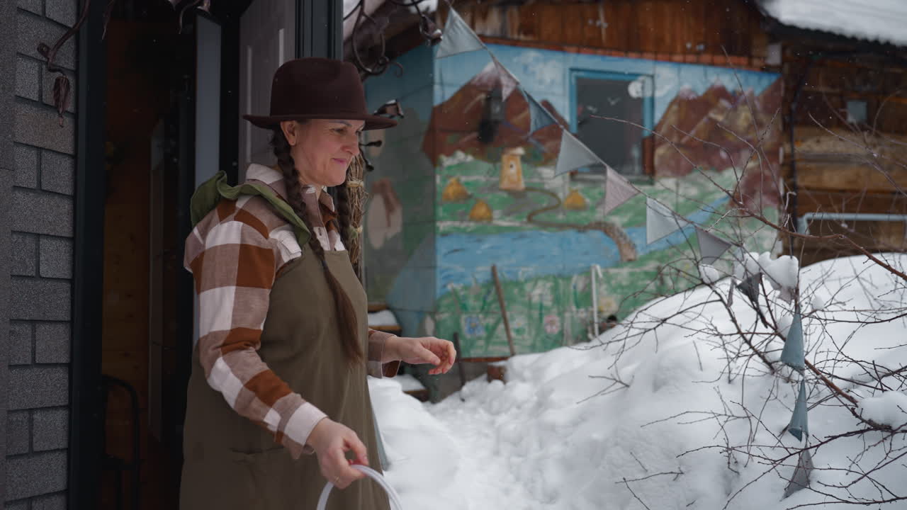 Homemaker wearing wide brim hat and olive gown steps into snowy yard holding empty bucket, scooping fresh snow with bucket and gloved hands while loyal dog follows against colorful mural backdrop