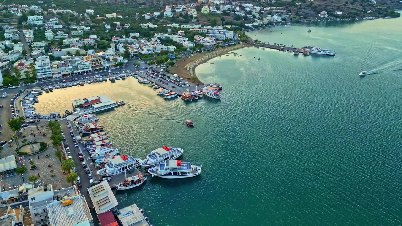 Turbåtar, parkerade bilar och en böjd sandstrand kantar Eloundas skyddade hamn medan kameran glider över vattenlinjen