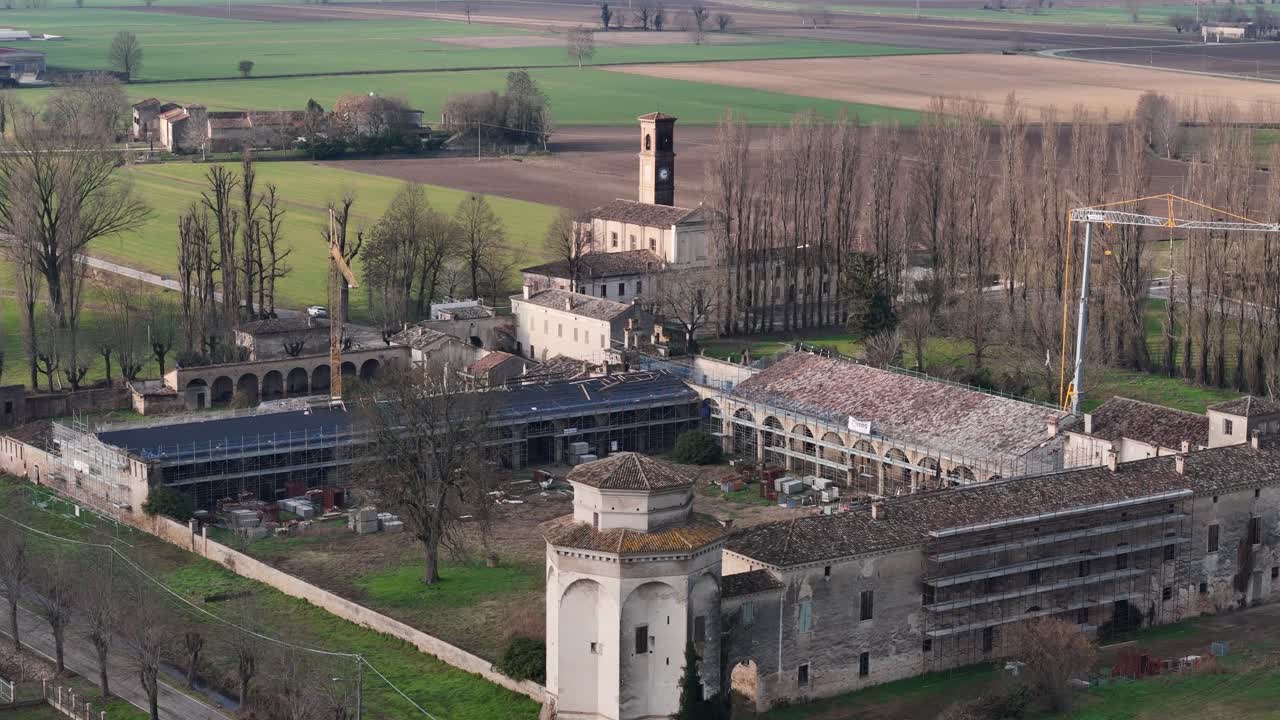 Letecký pohled na historický komplex venkovského dvorce Casatico Corte Castiglione, obklopený zemědělskou půdou Pádské nížiny, zachycující lešení, práce na rekonstrukci střechy, nádvoří, arkády a zimní stromy.