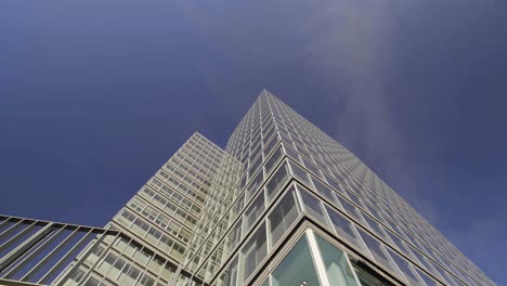 Tiro-Inclinado-Hacia-Arriba-Con-Giro-Lento-De-Una-Moderna-Torre-De-Oficinas-De-Vidrio-Y-Aluminio-En-Un-Día-Soleado-Con-Un-Cielo-Azul-Claro-Y-Un-Toque-De-Nubes