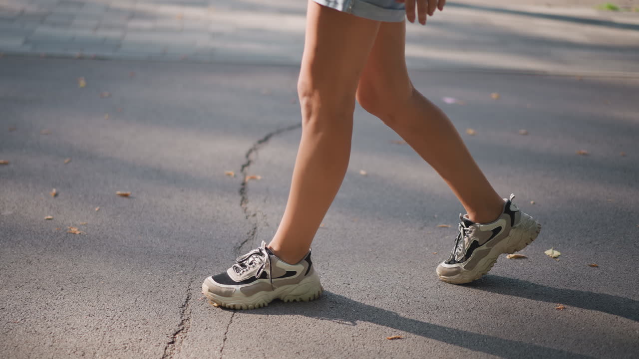 Donna bianca che cammina sul marciapiede nella morbida luce della sera, sneakers sull'asfalto, pantaloncini casual e passo calmo. Attenzione alla solitudine e al benessere, seguendo la propria filosofia di vita personale per il movimento quotidiano.