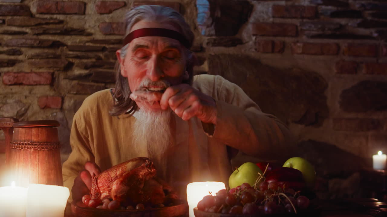 Elderly guest takes part at medieval party. Hungry mature bearded lord eats food and drinks wine at dining table in lord castle. Historical reconstruction