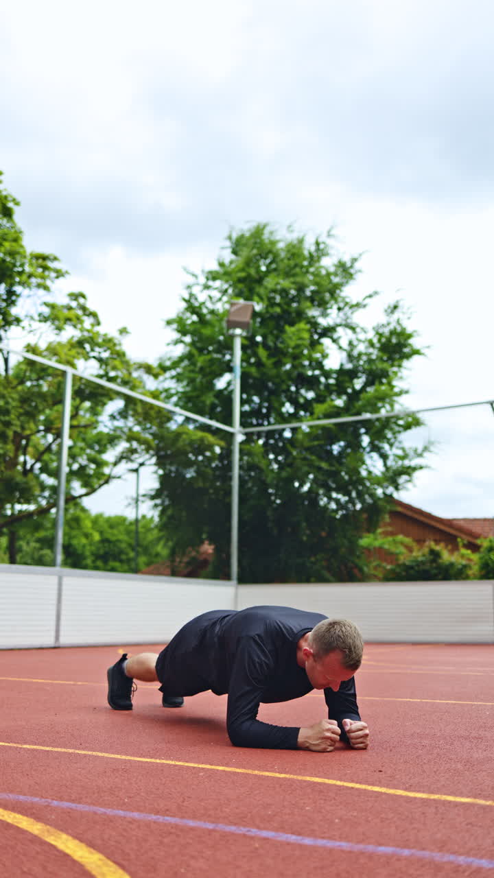 Uomo atletico che allunga le braccia alle sbarre parallele in una palestra all'aperto nel parco. Vestito con uniforme nera, pantaloncini scuri e scarpe da ginnastica. Si allena all'aperto.