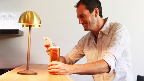 hombre sonriente usando un teléfono móvil mientras toma cerveza en el mostrador