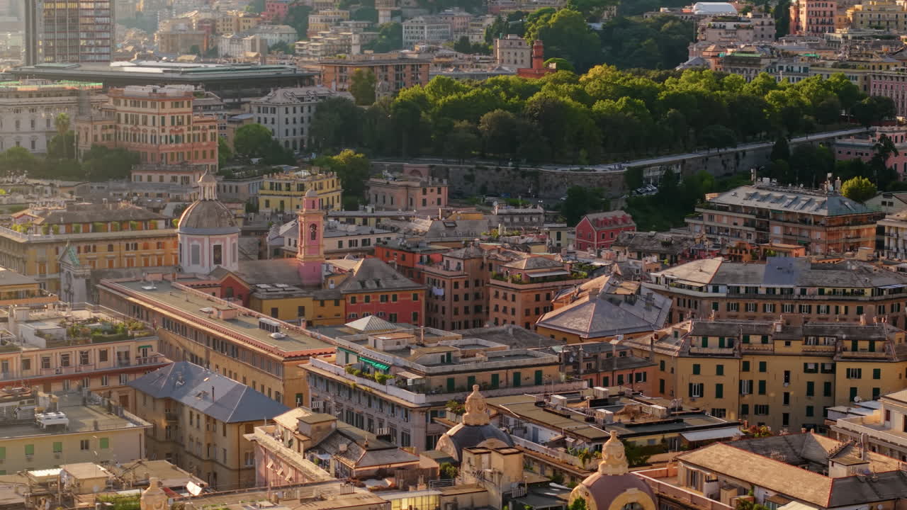 The drone advances with a panning motion above the old town of Genoa, showing rooftops, trees, and historic buildings at sunset