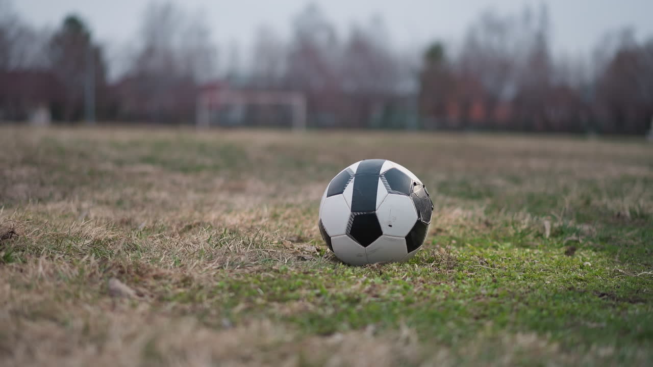 Close-up of two people approaching a soccer ball on a grassy field, the background is blurred, showcasing a football goalpost