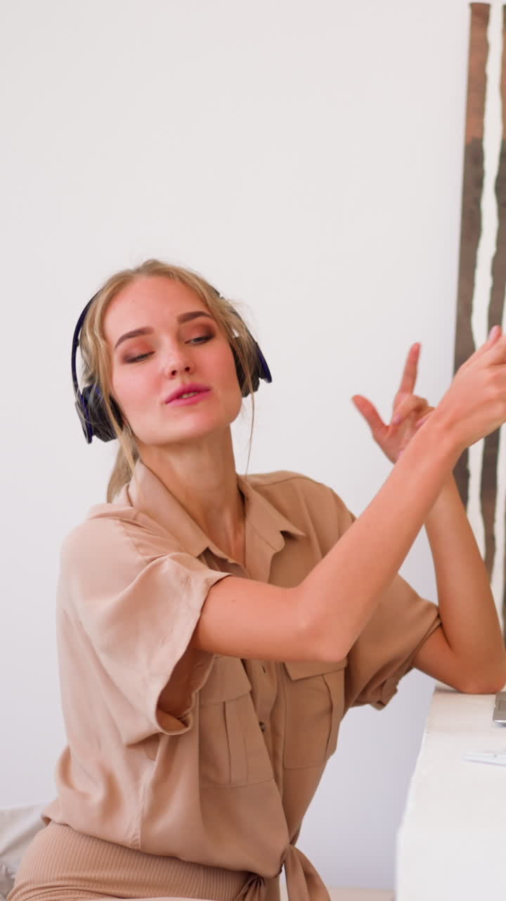Happy blonde woman with ponytail in headphones enjoys dancing music and throws paper sitting in front of modern laptop at white table slow motion