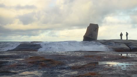 Zwei-Männer-Blicken-Aufs-Meer-Hinaus,-Während-Wasser-Aus-Dem-Granit-Blasloch-Schießt