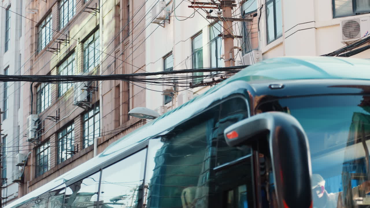 Autobús urbano enmarcado por cables, un autocar turquesa de movimiento lento pasa por una avenida estrecha bordeada de fachadas de mediados de siglo, cables aéreos enredados, semáforo y ventanas reflectantes crean una escena urbana cinética.