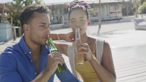 Happy-diverse-couple-talking-and-drinking-beers-on-beach-sun-deck,-in-slow-motion