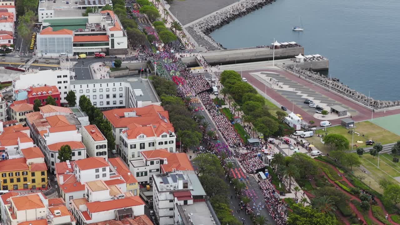 Aerial orbit shot of Funchal, Madeira during the vibrant Flower Festival, capturing colorful floats, crowds, and festive atmosphere along the scenic coastal promenade