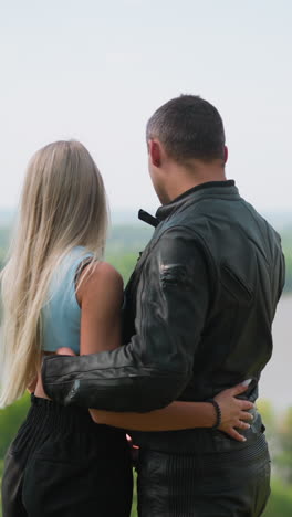 long haired blonde woman and motorcyclist in protective suit hug looking at tranquil river from high hill slope on spring day backside view slow motion