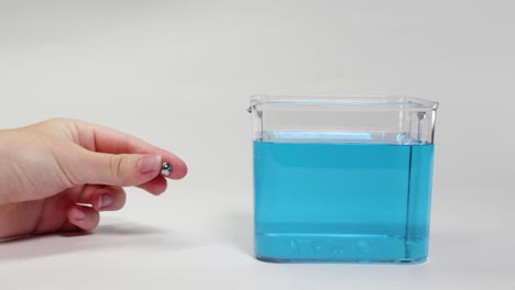 a hand drops a metal ball bearing into a clear container filled with blue liquid, observing its descent