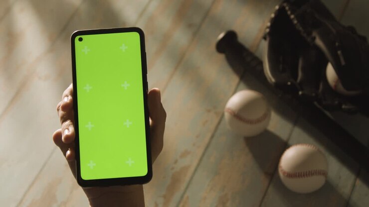 Persona con cellulare a schermo verde sopra una natura morta di baseball con mazza, palla e guanto da ricevitore su pavimento in legno invecchiato