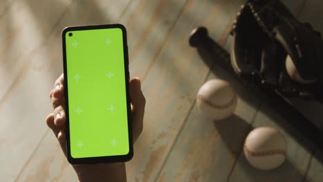 Person-Holding-Green-Screen-Mobile-Phone-Above-Baseball-Still-Life-With-Bat-Ball-And-Catchers-Mitt-On-Aged-Wooden-Floor