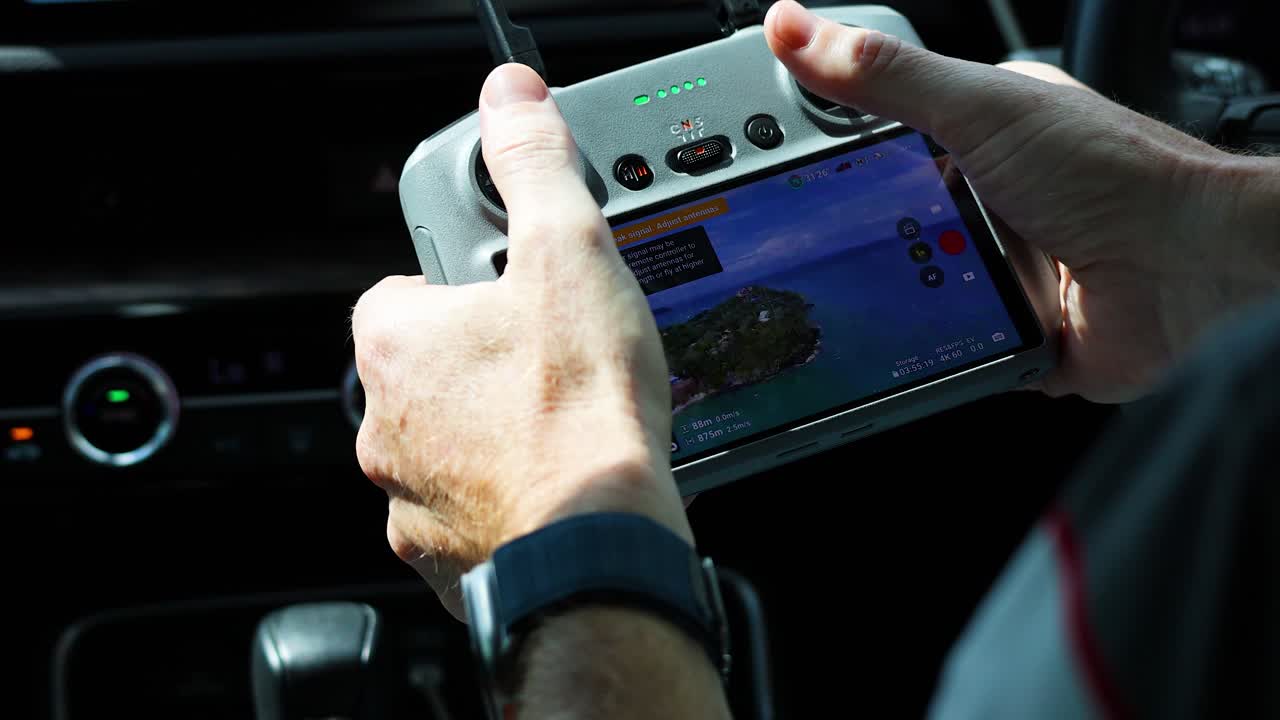 Hands operate a drone controller inside a vehicle, displaying a scenic view on the screen. Bright lighting enhances focus