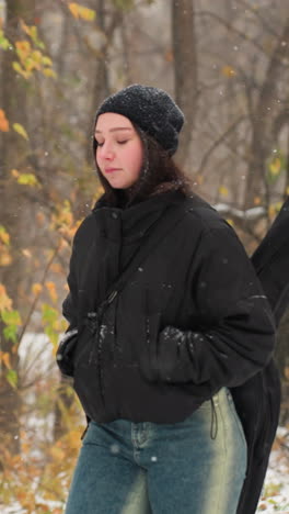 joven con chaqueta de invierno con las manos en los bolsillos y la guitarra en la espalda caminando en la nieve, banco de parque nevado y coloridas hojas de otoño en el fondo