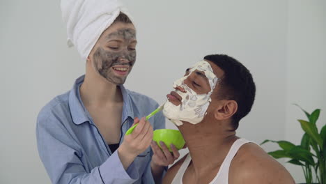 mujer con máscara facial aplicando exfoliante a un apuesto hombre negro