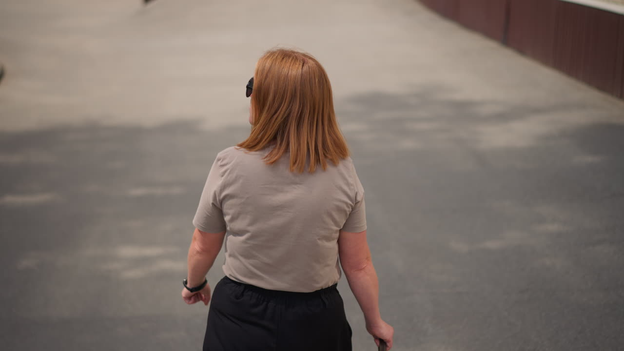 Rear view of business lady with light brown hair walking along pavement toward school building under warm sunlight, checking wrist watch and looking left and right