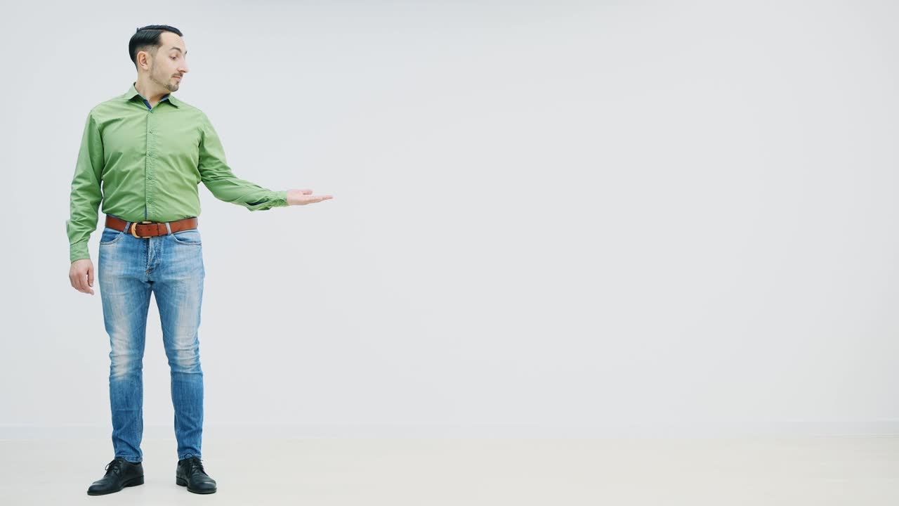 Full-length young handsome casual man appearing on the white background, showing at something, giving thumb up, looking to the camera. Funny face expression.