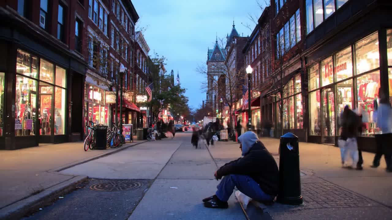 As twilight settles, people walk through a vibrant urban street filled with shops. A lone figure sits on the curb, highlighting the contrast between bustling activity and solitude.