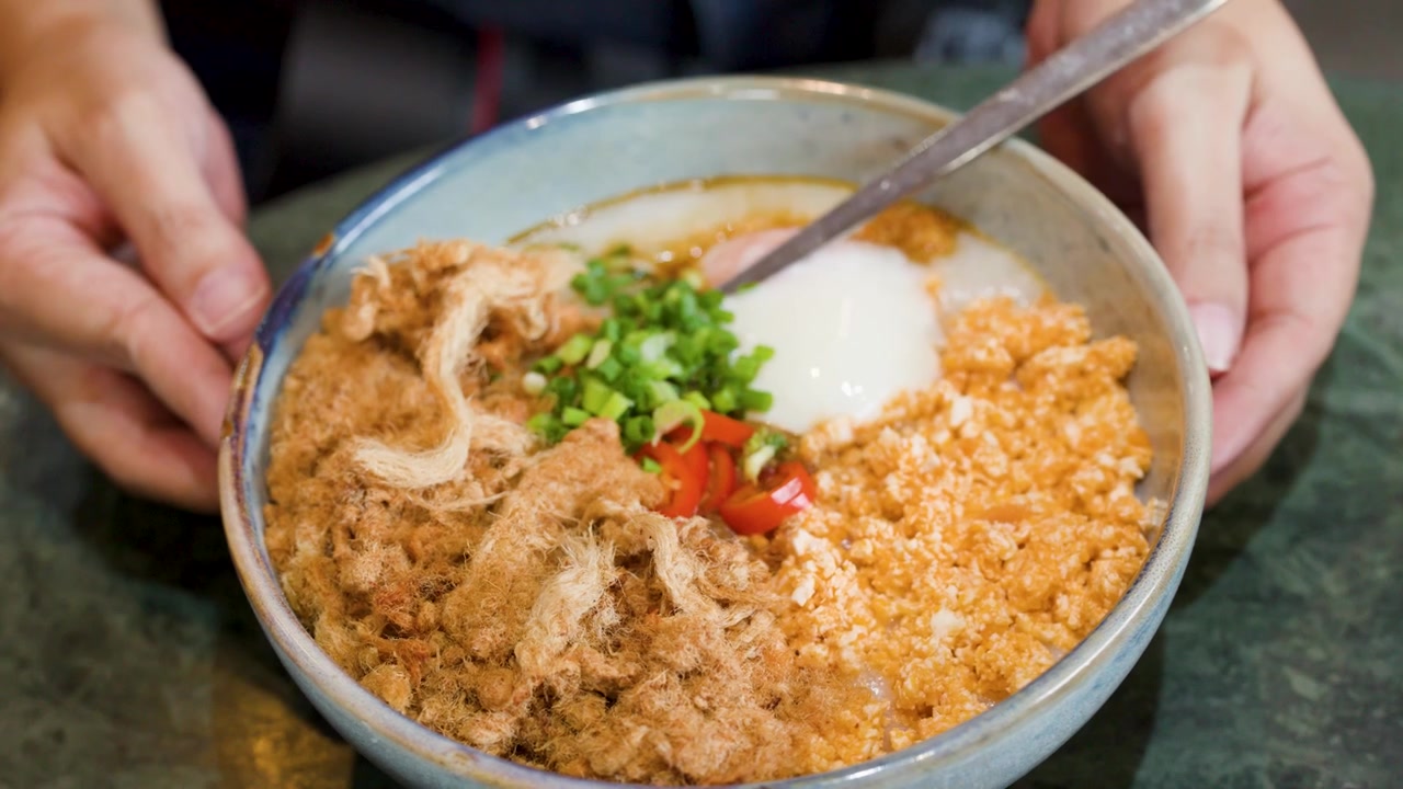 Cận cảnh đôi tay xoay một bát cháo gạo mặn với các loại topping truyền thống