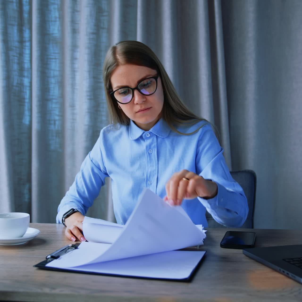 Busy lady wearing glasses working in office at her desk. Employee looks through the documents quickly, turning pages. Grey backdrop