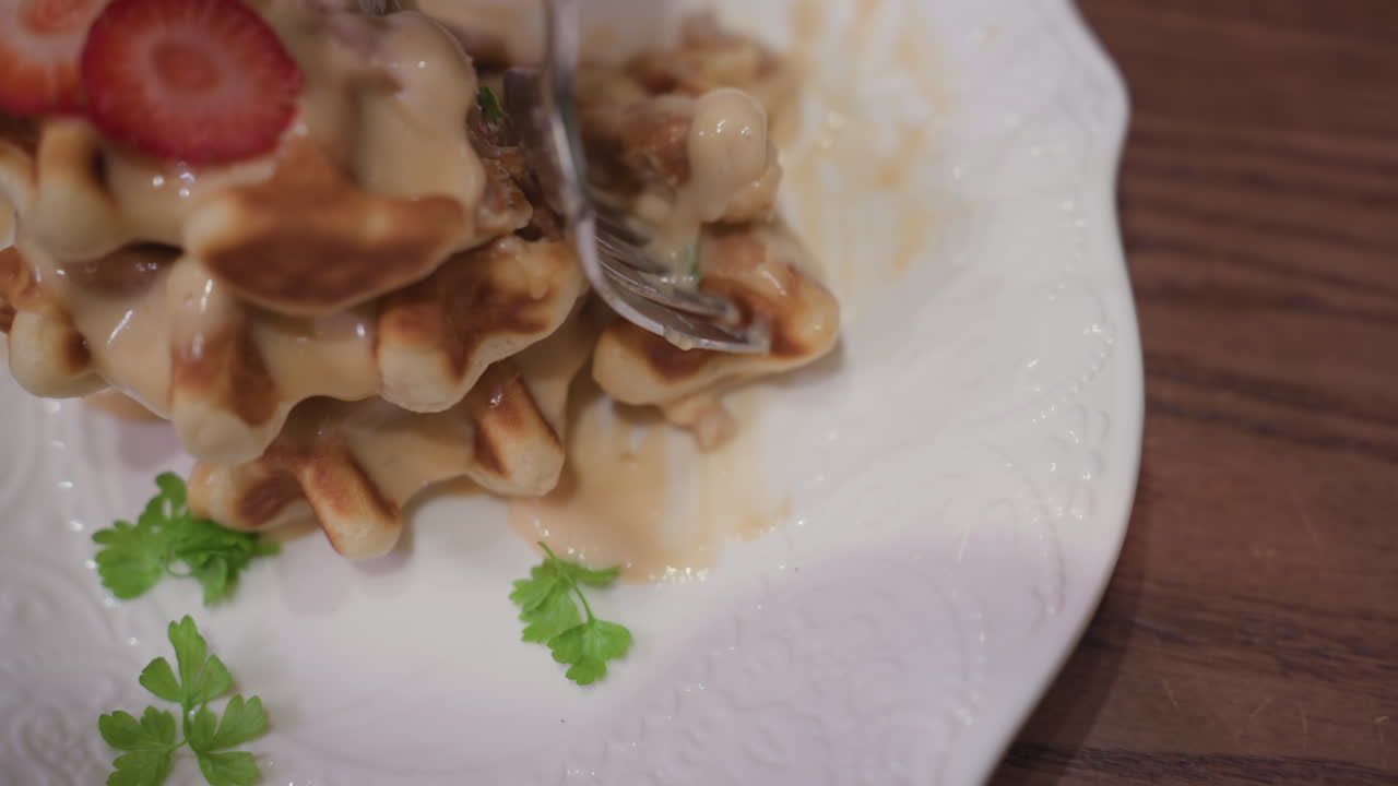 Close up of fork holding waffle piece dipped into smooth creamy sauce on elegant white plate, topped with fresh strawberry slices and garnished with parsley leaves, set on wooden tabletop surface