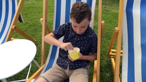 A-smartly-dressed-boy-eats-ice-cream-from-a-tub,-sitting-on-a-deck-chair