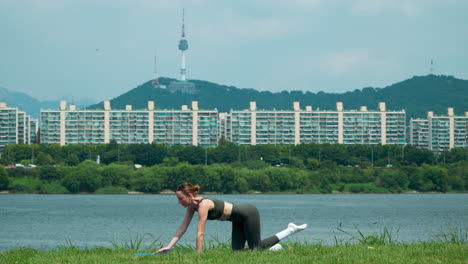 Mujer-Atleta-Haciendo-Ejercicio-De-Tabla-De-Perro-Pájaro-En-El-Parque-Del-Río-Han-Sobre-Césped-Con-Vistas-A-La-Torre-Namsan-De-Seúl