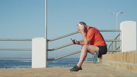 Hombre-Mayor-Usando-Auriculares-Usando-Un-Teléfono-Inteligente-En-El-Paseo-Marítimo