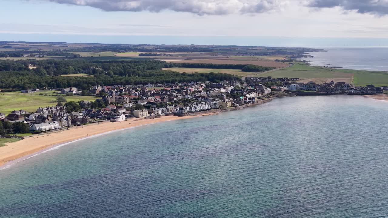 Drone camera smoothly pans above seaside town, sandy beach, and bay under partly cloudy daylight