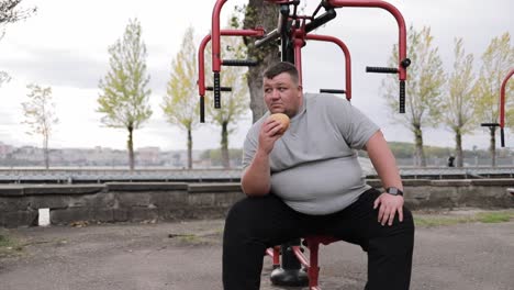 a very fat man sits on an outdoor exercise machine and holds a bun a very fat man sits on an outdoor exercise machine and holds a bun a quick and unhealthy snack struggle with excess weight