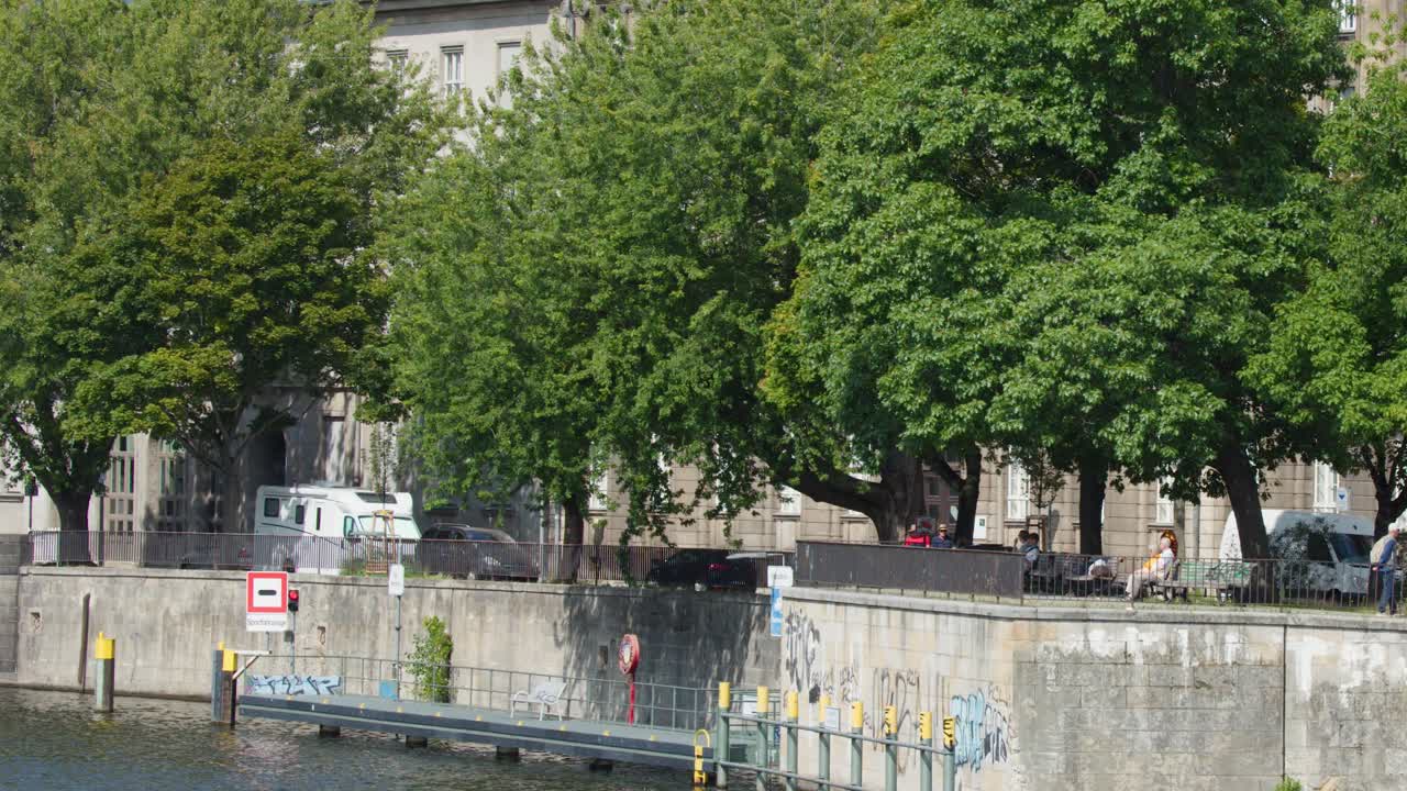 People stroll beside Spree River, leafy trees, historic architecture, daylight, static wide shot