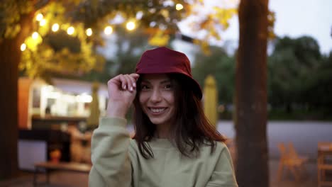 a young brunette puts on a red stylish hat against the background of yellow coffee lanterns. and evening walk