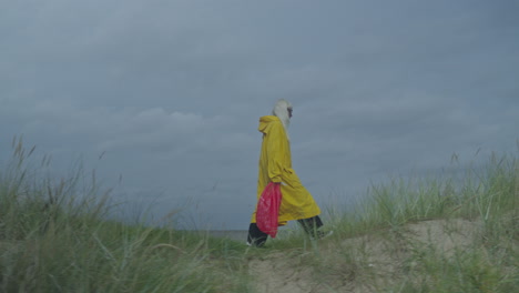 Mujer-Con-Impermeable-Amarillo-Y-Bolsa-Roja-Caminando-Por-Una-Duna-De-Arena,-Capturada-Con-Una-Cámara-Lateral