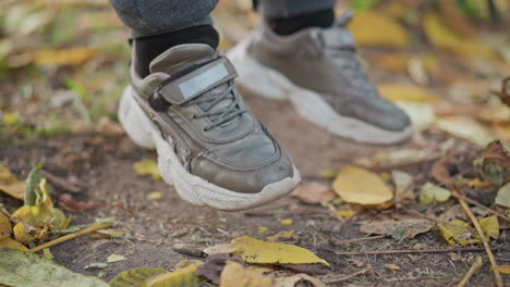 close up of child legs in grey joggers and chunky sneakers hovering above autumn leaves and pine needles on park ground with leaves gently swaying in breeze rustic fall setting with seasonal motion