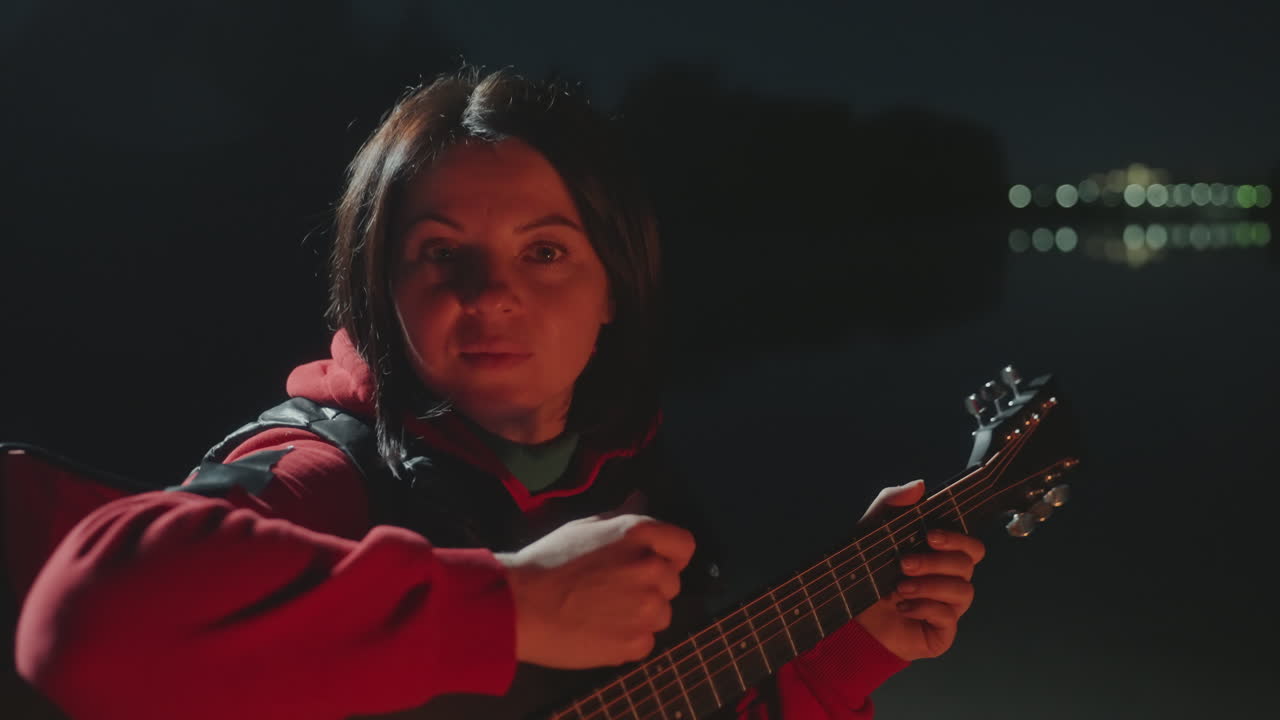 Donna bianca in campeggio in riva al lago, chitarra di notte accanto al falò, sedia pieghevole rossa, caldo bagliore del fuoco, luci della città in lontananza sull'acqua, espressione contemplativa, maglione accogliente, morbido strimpellare, fumo che si disperde.