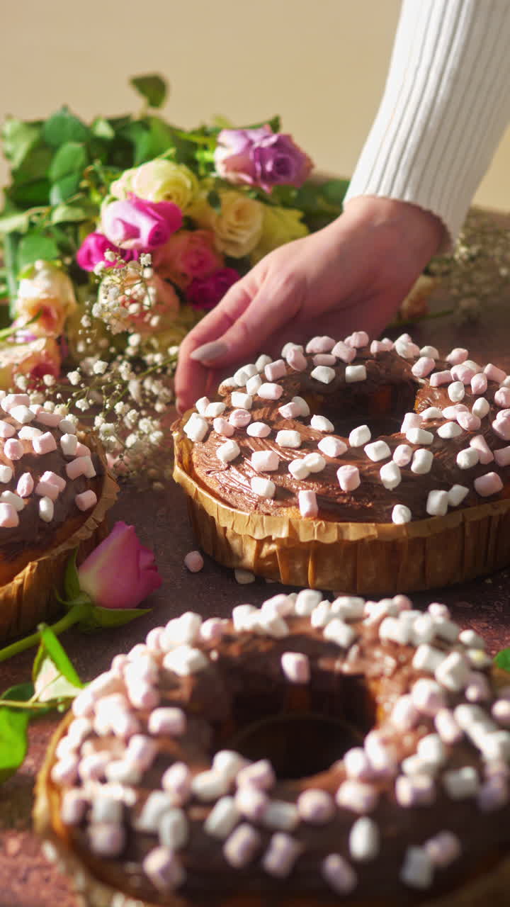 Hand placing donut shaped chocolate cake topped with mini marshmallows on a decorated dessert table with roses and sweets. Festive bakery scene for birthday and Valentine’s Day