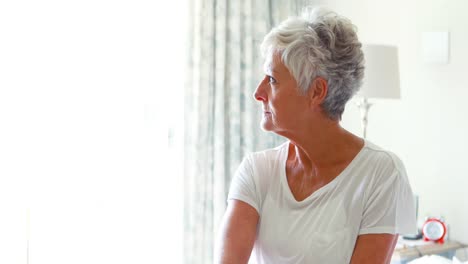 Thoughtful-senior-woman-sitting-on-bed-in-bedroom