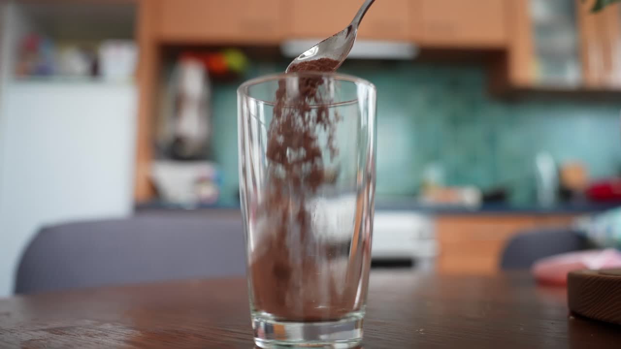 Un cucchiaio versa cacao in polvere in un bicchiere alto su un tavolo di legno, primo piano con sfondo sfocato di una cucina, per preparare una cioccolata calda fatta in casa